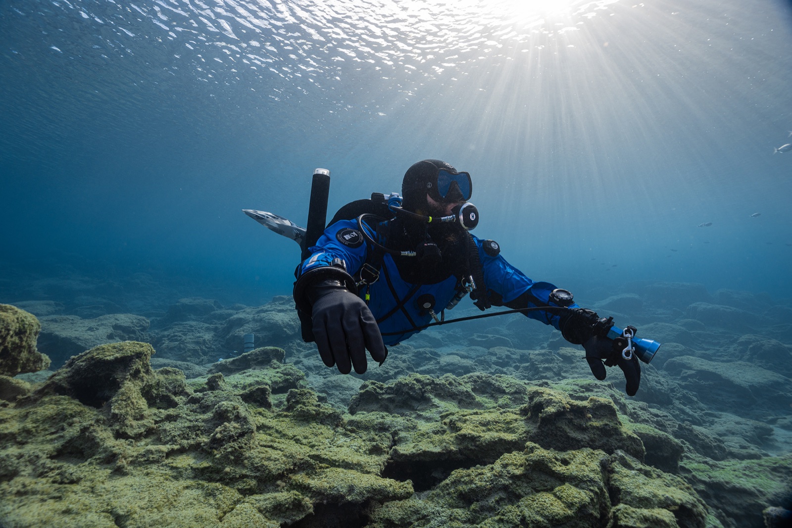 GUE tech diver in blue drysuit gliding over Mediterranean rocky reef with sun rays